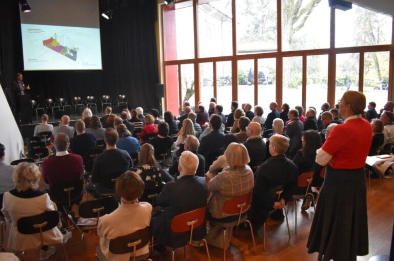 Ein großer Konferenzraum mit vielen Menschen die auf eine Leinwand schauen.